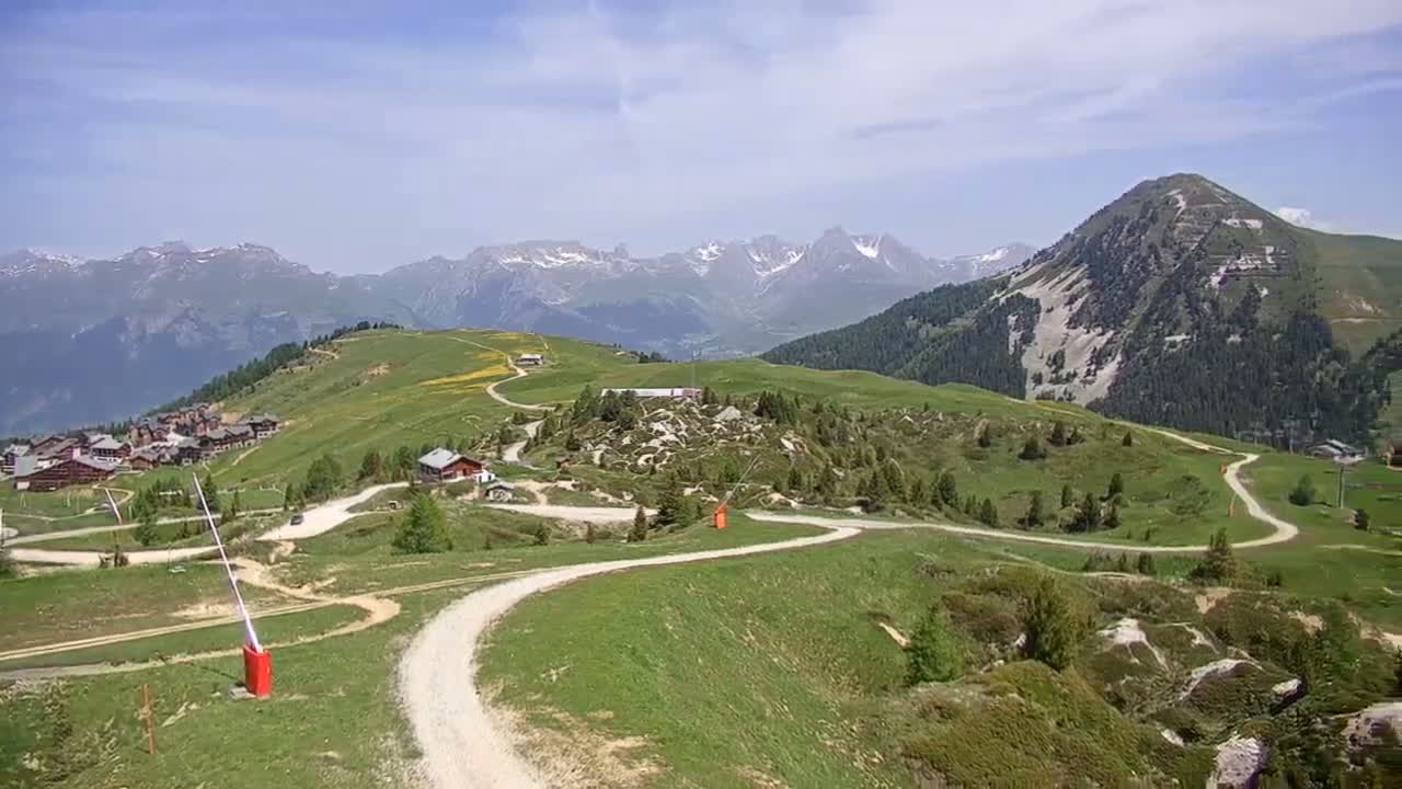 A winding dirt road meanders through a grassy mountain landscape under a mostly sunny sky, with a small village and snow-capped mountains in the distance.