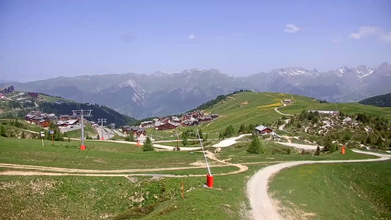 A mountain village nestled in a green valley, overlooked by a snow-capped mountain range under a clear blue sky.