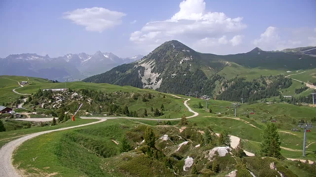 A winding dirt road snakes through rolling green hills and sparsely wooded mountains under a partly cloudy sky.