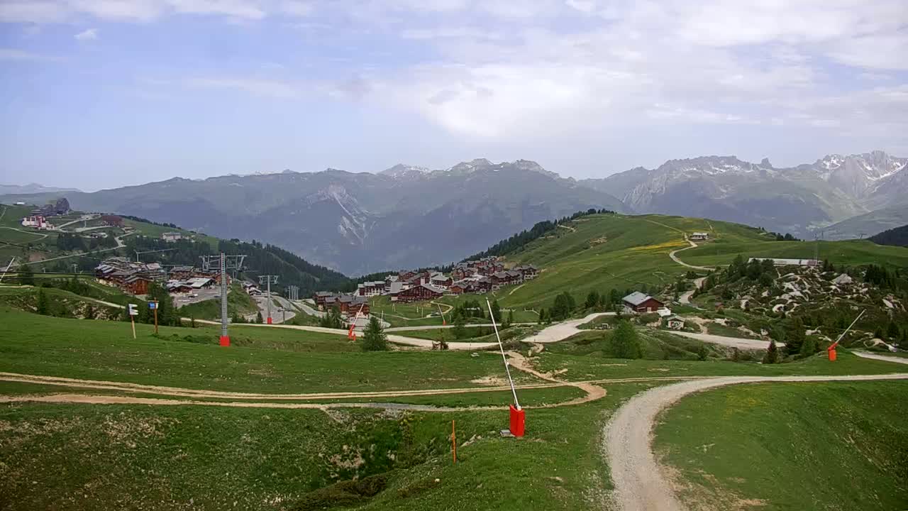 A mountain village nestled in a green valley under a partly cloudy sky, with ski lifts and winding roads visible.