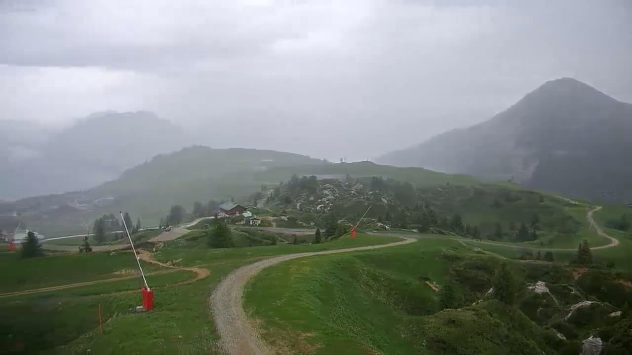 A winding dirt road curves through a rain-swept, grassy mountain landscape, with hazy mountains in the background.