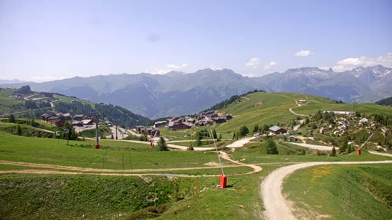 A mountain village nestled in a green valley, under a clear blue sky, with distant mountain peaks in the background.