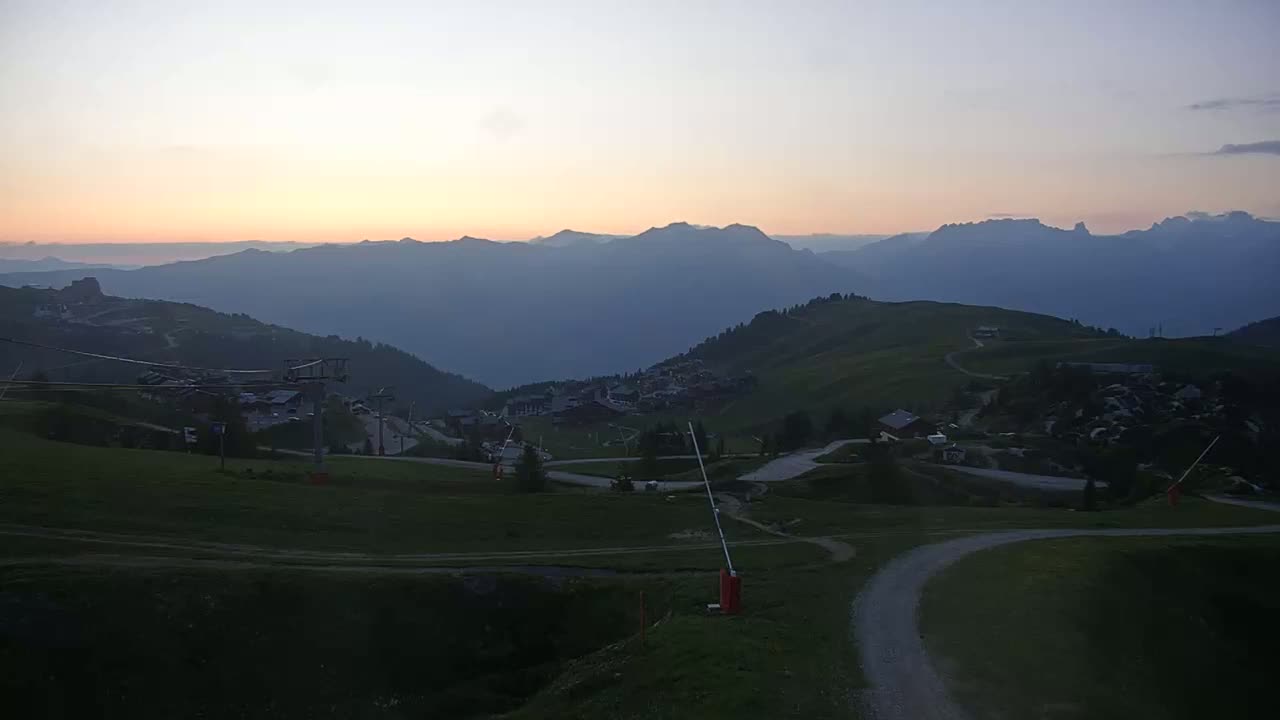 A mountain village nestled in a valley at sunset, with hazy mountains in the background and a ski lift visible in the foreground.