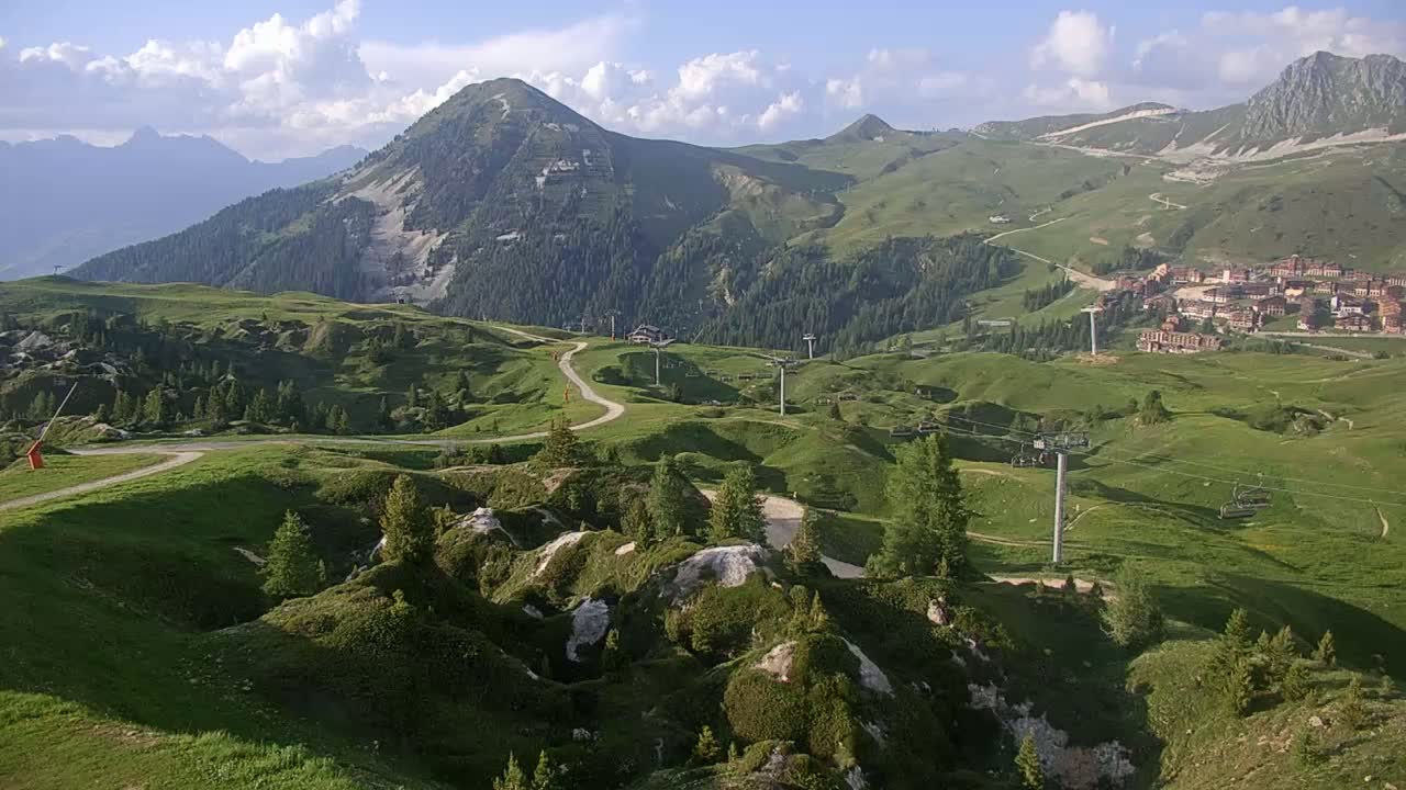 A partly cloudy day reveals a mountainous landscape with rolling green hills, ski lifts, and a small village nestled in the valley.