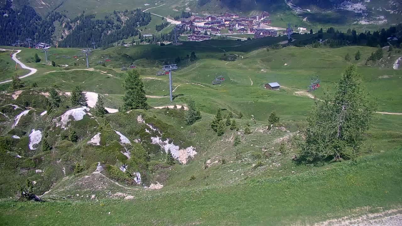 A high-angle view shows a sunny, mountainous landscape with green hills, ski lifts, and a small village in the distance.