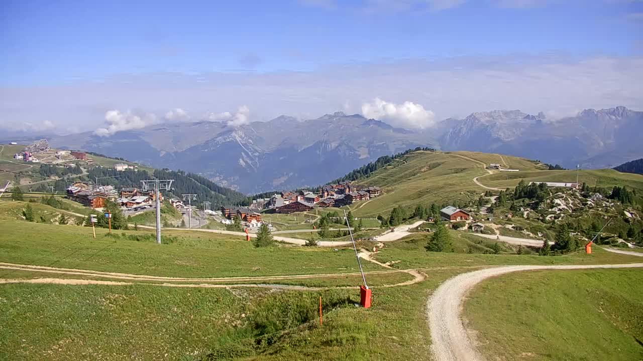 A mountain village nestled in a valley, with rolling green hills and a clear blue sky dotted with a few clouds.