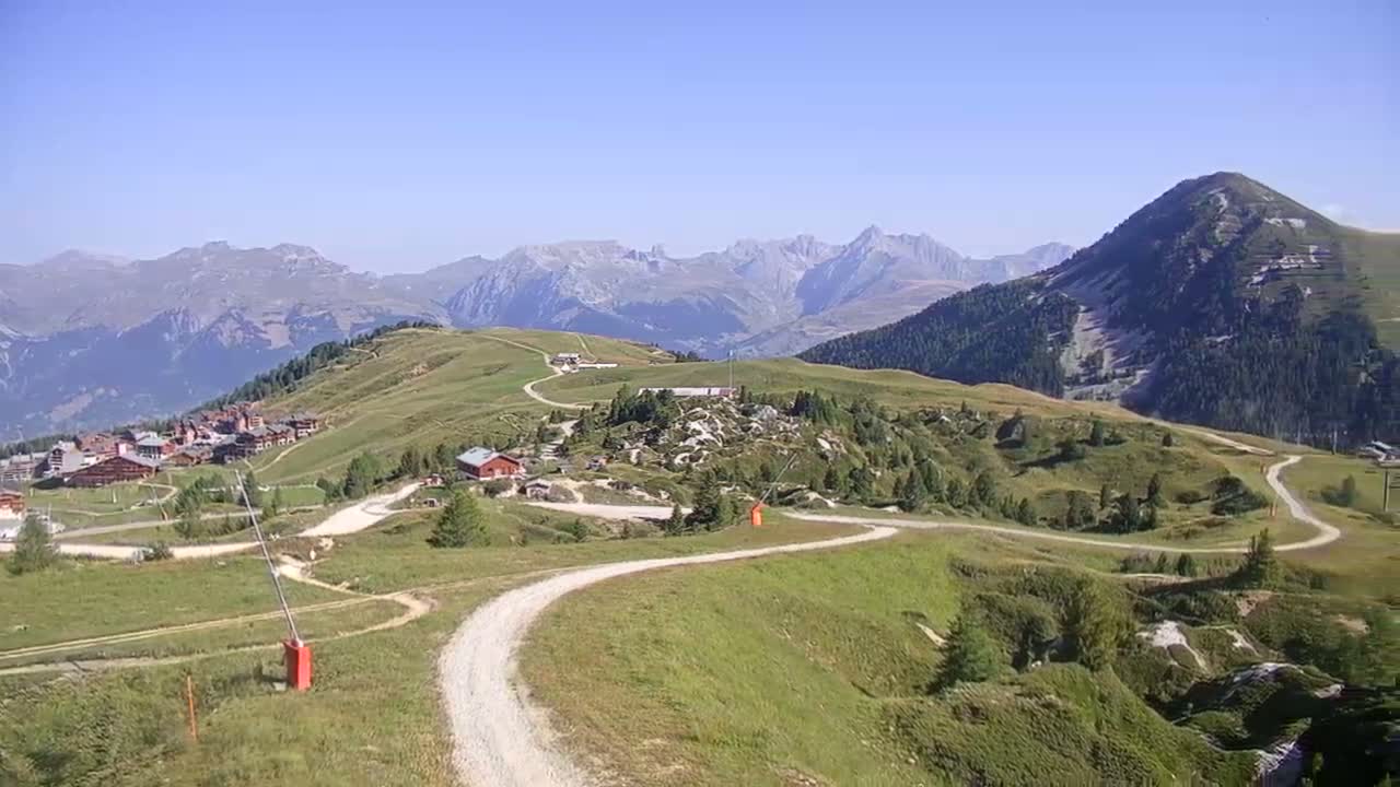 A winding dirt road leads through green rolling hills toward a village nestled in a valley, with a mountain range visible in the background under a clear blue sky.