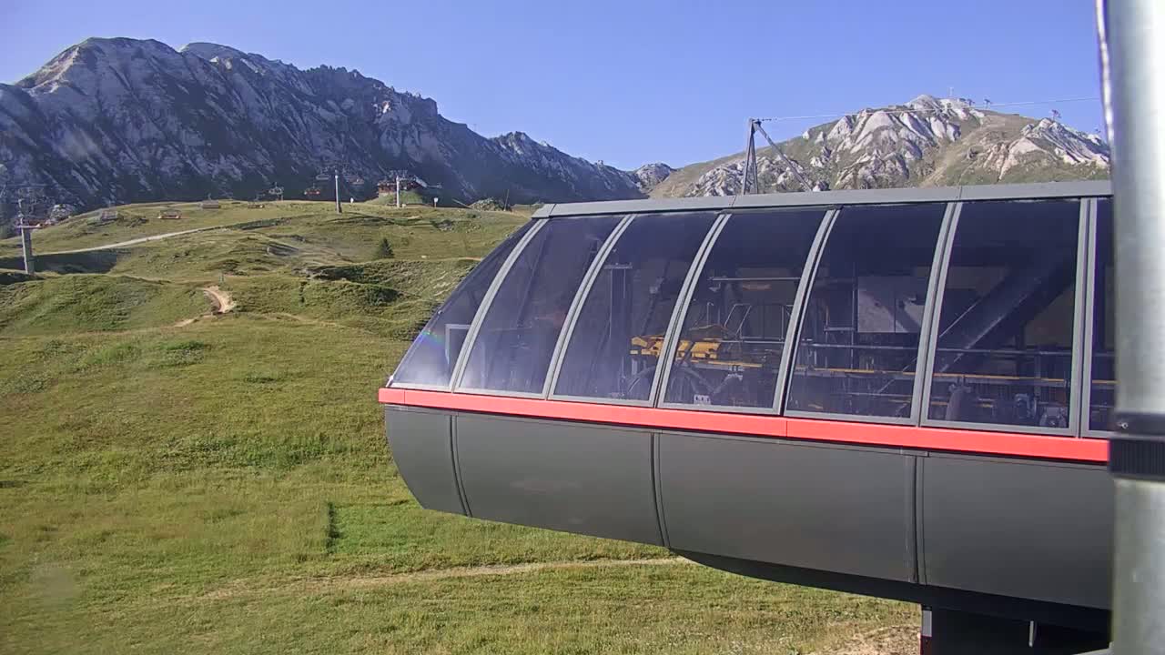 A portion of a gondola car is visible in the foreground, overlooking a grassy mountainside with distant, rocky peaks under a clear, sunny sky.