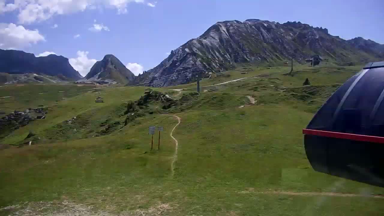 A portion of a gondola car is visible in the foreground, overlooking a grassy mountainside with distant, rocky peaks under a clear, sunny sky.
