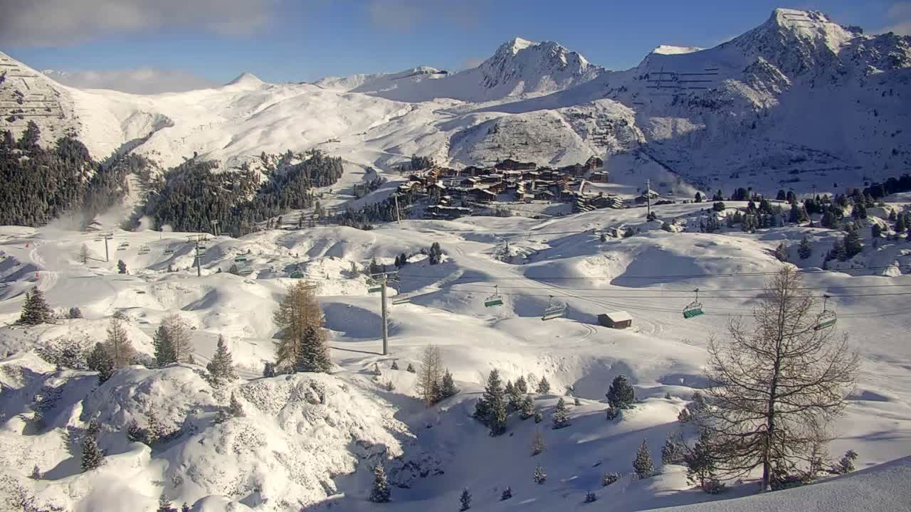 A bright, clear winter day reveals a vast snow-covered mountain landscape featuring a ski resort village nestled among slopes, numerous chairlifts, and scattered trees.