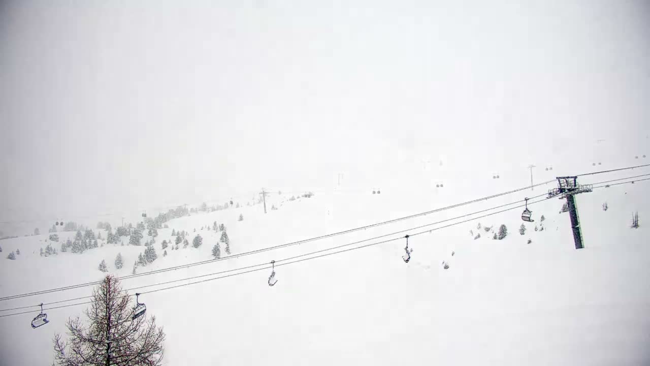 Empty ski lift chairs traverse a snow-covered mountain slope dotted with evergreen and bare trees, under heavy overcast conditions with very low visibility from snow or fog.
