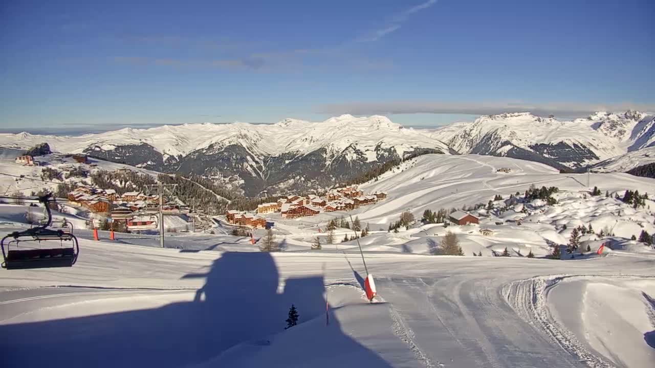 The image captures a sunny winter day at a snow-covered mountain ski resort, featuring a ski slope with a lift chair and shadows in the foreground, a village of chalets nestled in the valley, and a backdrop of majestic snow-capped peaks under a clear blue sky.