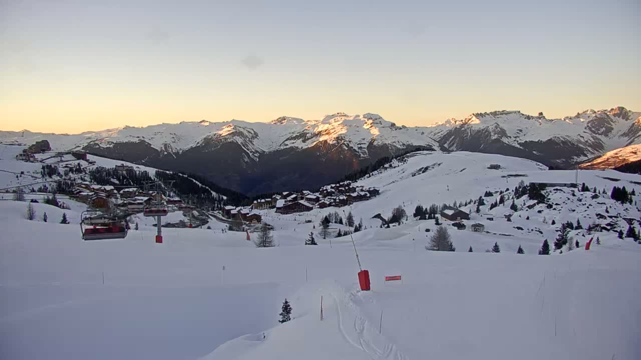 A sprawling snow-covered mountain landscape, featuring evergreen trees, multiple ski lifts, and a prominent peak with avalanche barriers, extends under a partly cloudy sky with patches of blue.