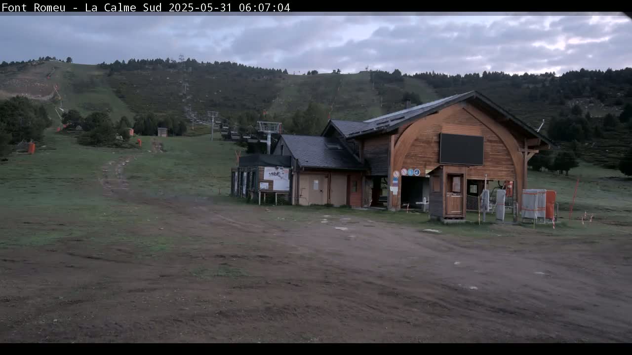 A wooden ski lift building sits in a grassy area with a hill covered in ski lifts in the background under an overcast sky.