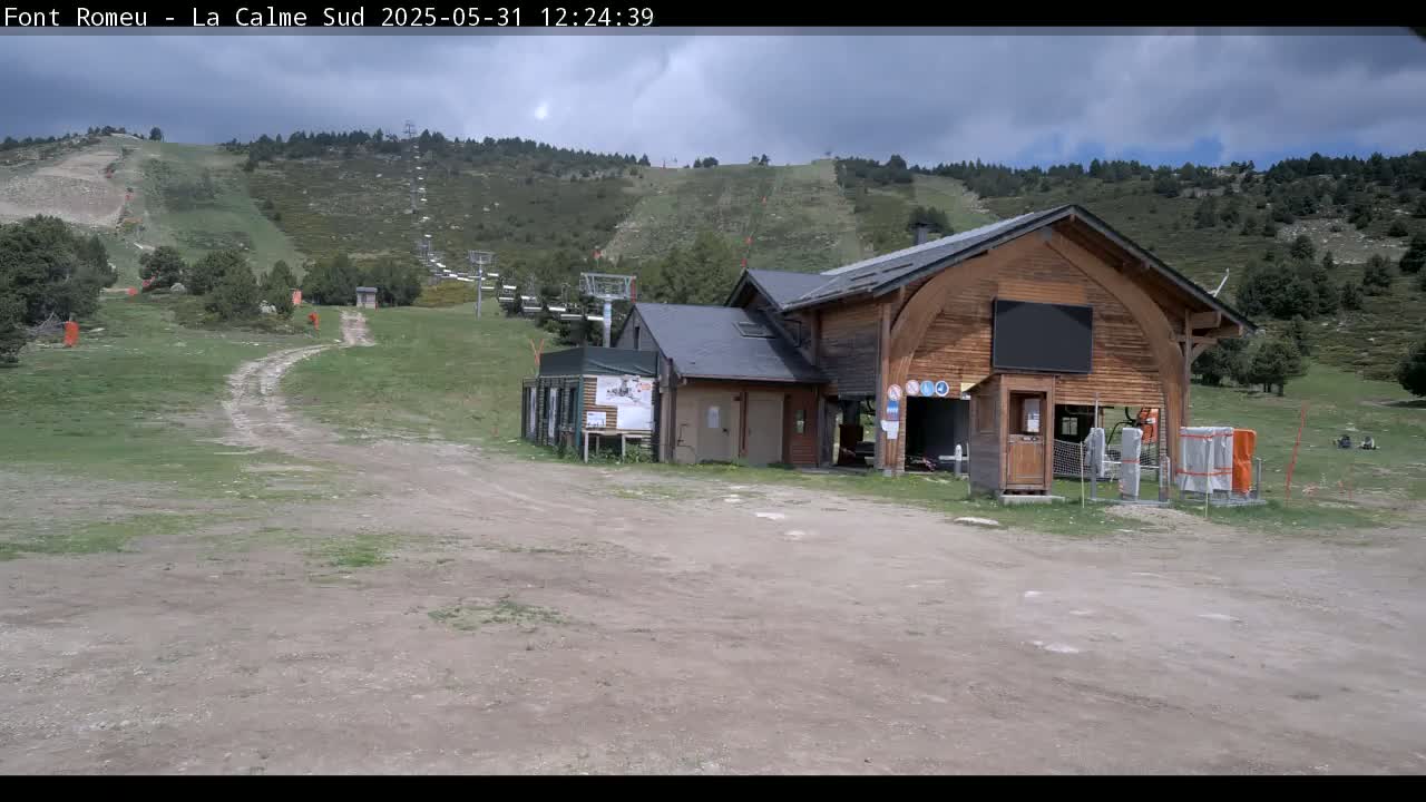 A wooden building sits in a grassy area near a ski lift on a partly cloudy day.