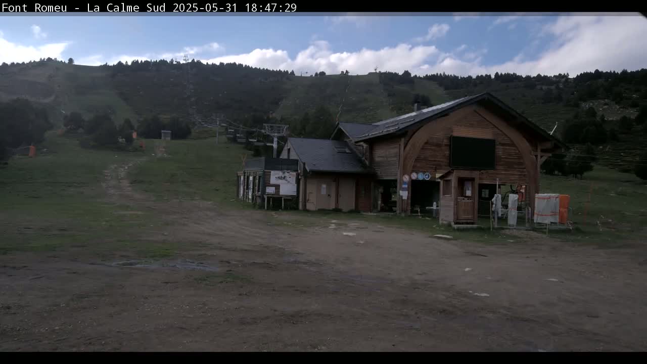 A wooden building sits in a grassy area near a ski lift against a hillside of dark green trees under a partly cloudy sky.