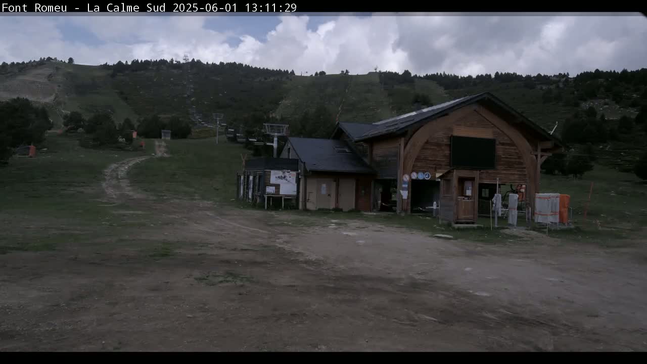 A wooden building sits near a ski lift at the base of a mountain on a cloudy day.