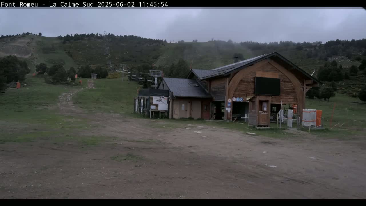 A wooden building sits in a grassy area with a ski lift ascending a hill in the background under an overcast sky.