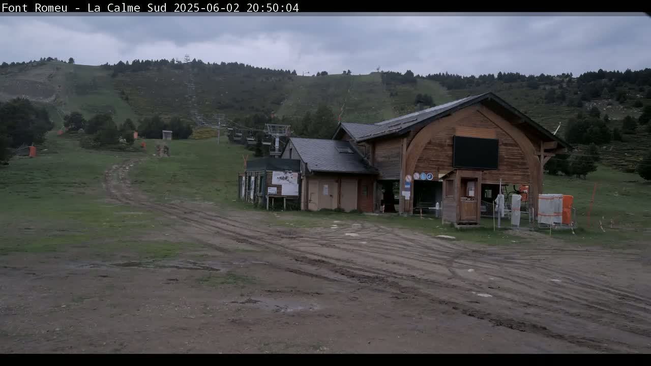 A muddy, grassy area with tire tracks leads to a wooden building and a ski lift against a backdrop of overcast hills.