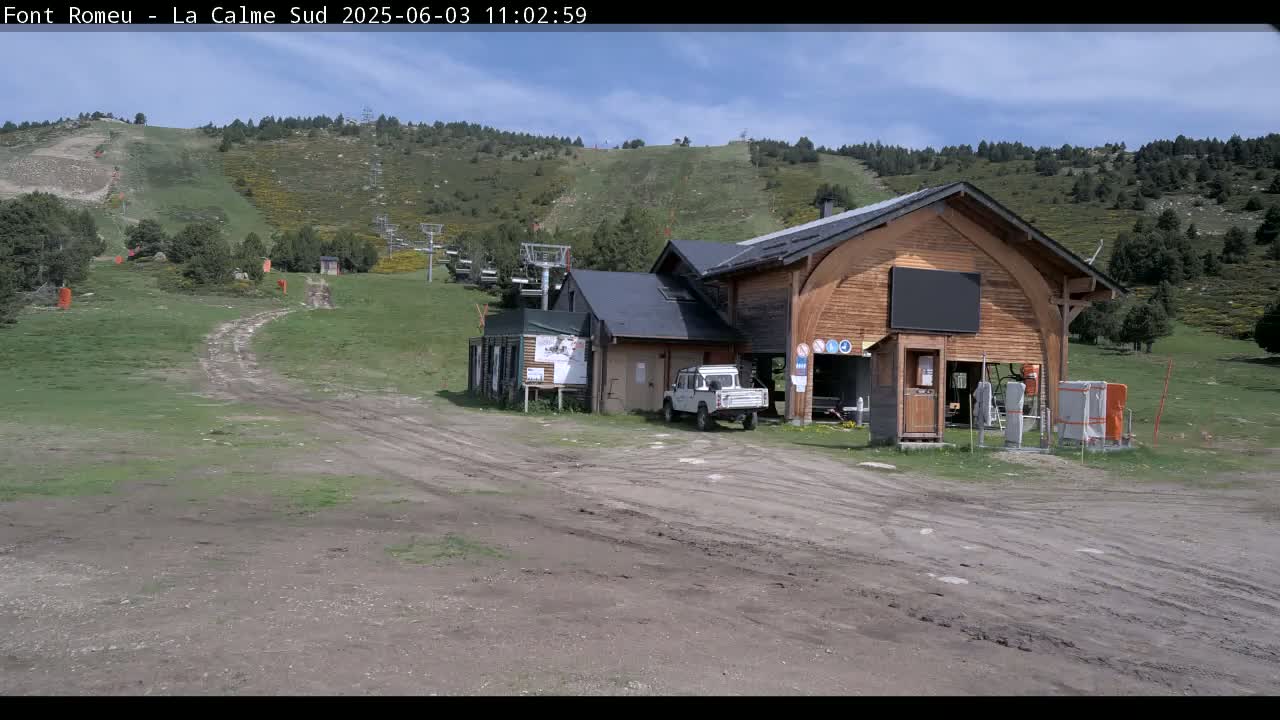 A mostly sunny day reveals a wooden building near a ski lift on a grassy hillside with a dirt road leading to it.
