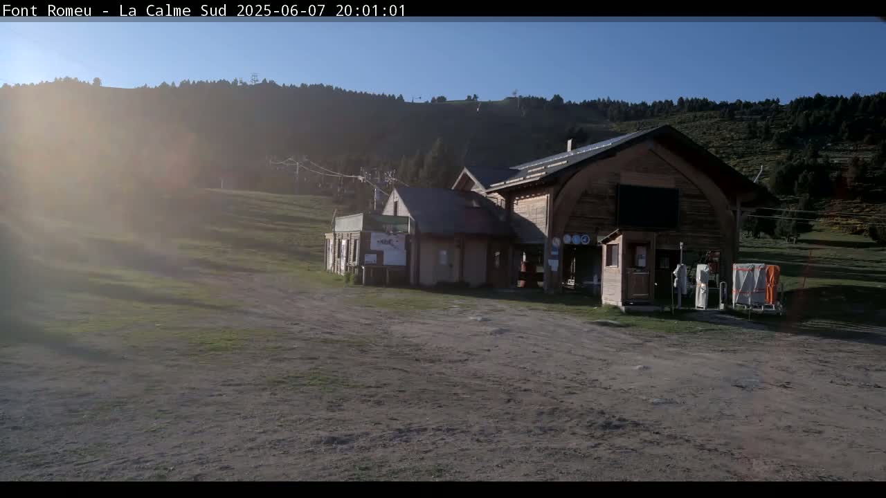 A brown wooden building sits in a grassy area, with a hillside covered in trees visible in the background under a clear, sunny sky.