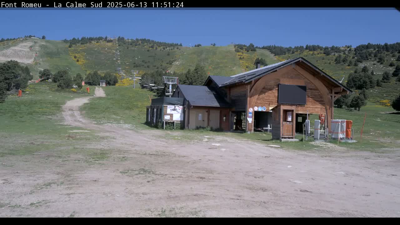 A wooden building sits near a grassy hillside with a chairlift and a dirt path under a clear blue sky.