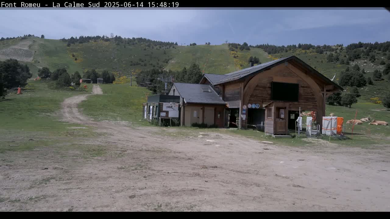 A wooden building sits in a grassy area near a dirt road leading up a hillside under a mostly clear, sunny sky.