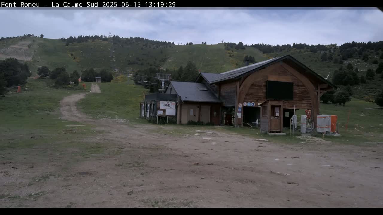 A wooden building sits near a grassy hillside with a dirt road leading up to a ski lift under a cloudy sky.