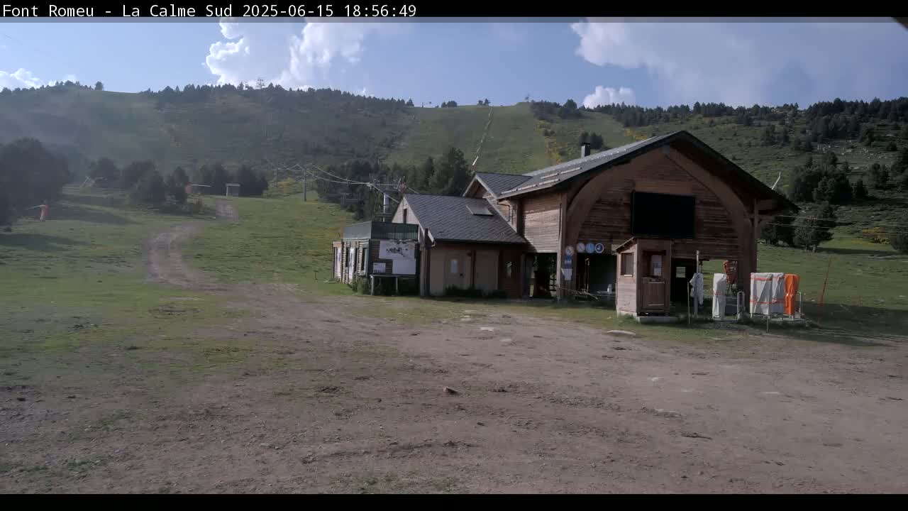 A wooden building sits in a grassy area near a ski lift against a backdrop of a partly cloudy sky and green hills.