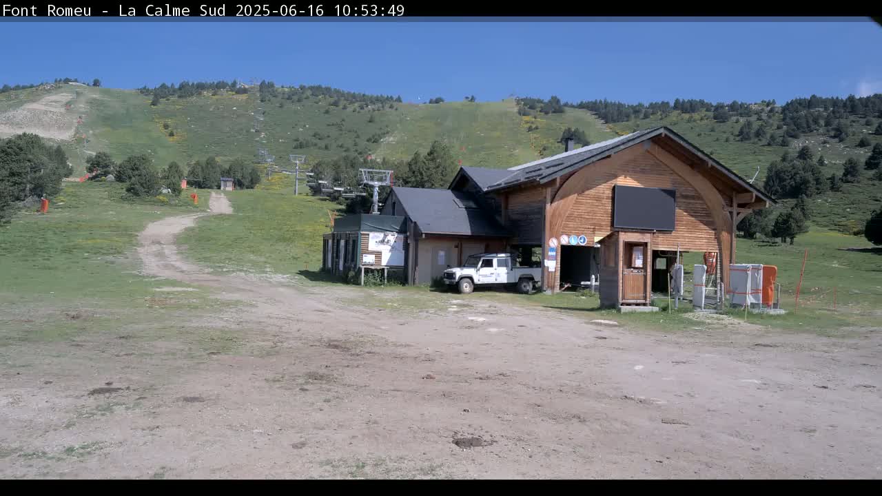 A grassy hillside with a ski lift and a large wooden building, a smaller adjacent building, and a white vehicle parked in front, under a clear blue sky.