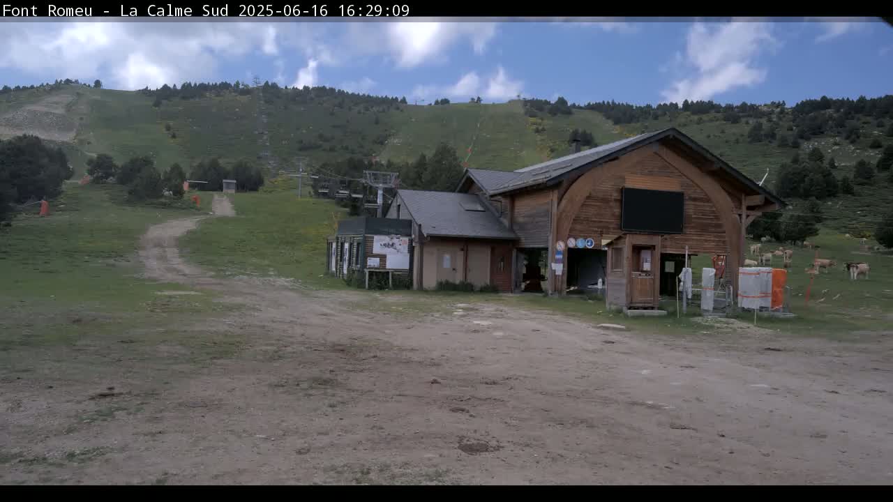 A dirt road leads to a wooden building and a grassy hillside with a ski lift in the background under a partly cloudy sky.