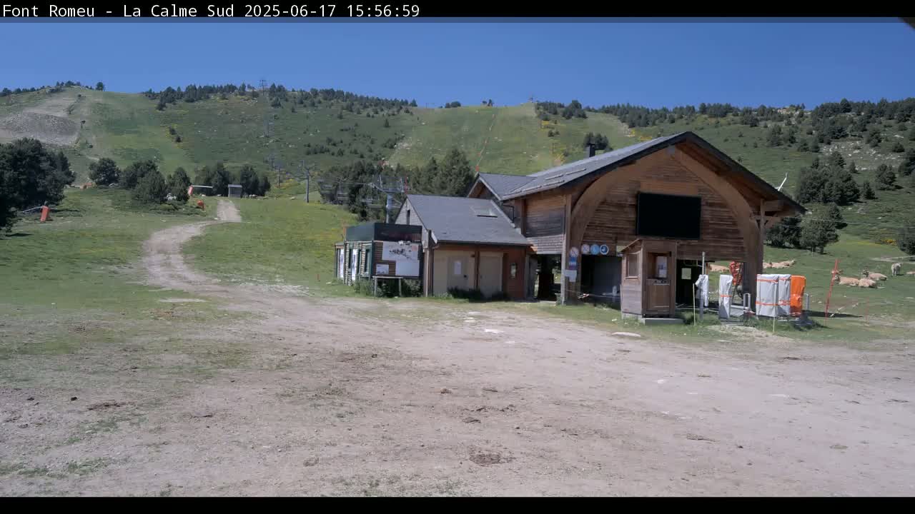 A wooden building sits near a grassy hill on a sunny day, with a dirt path leading up to it.