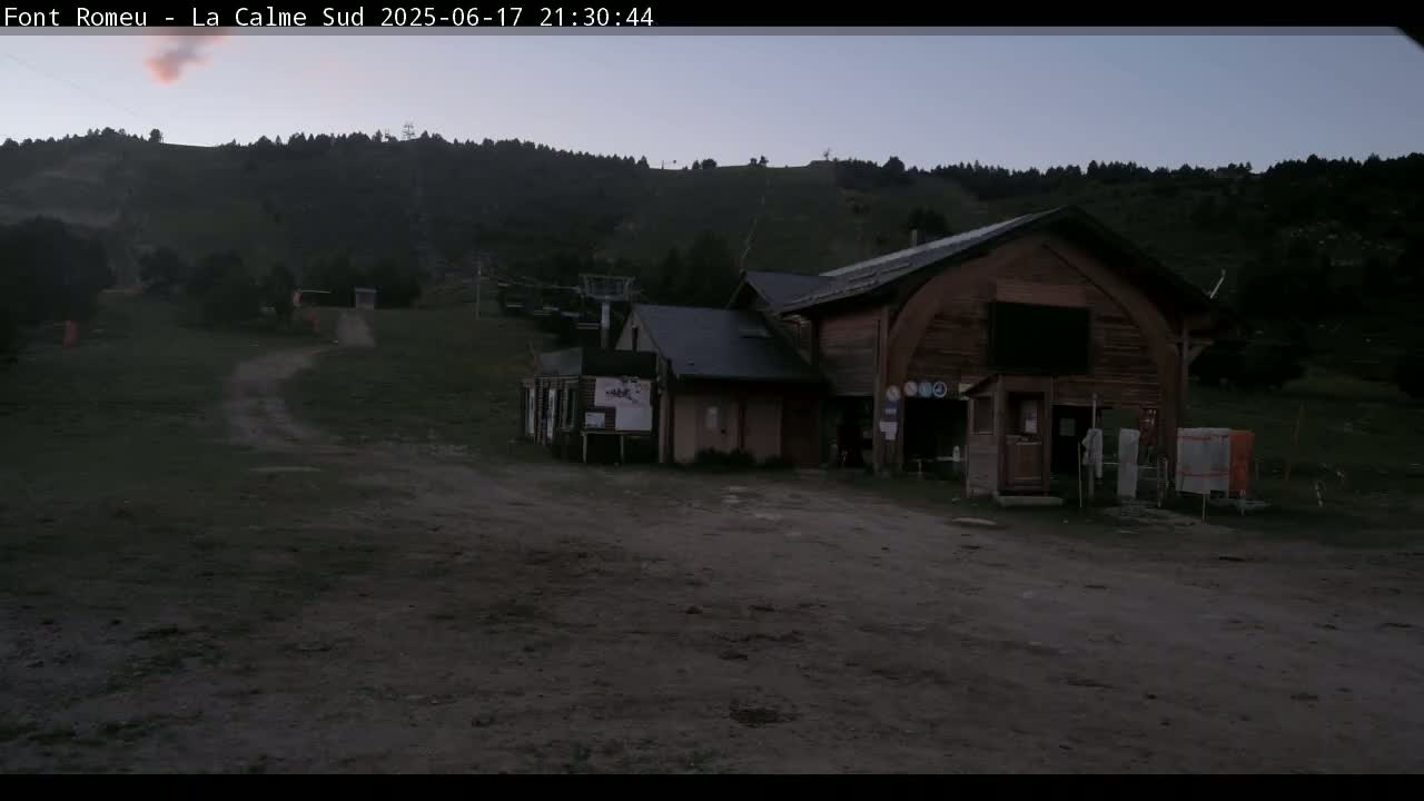 A dark brown wooden building sits in a dirt lot at the base of a hill covered in dark green trees under a twilight sky.
