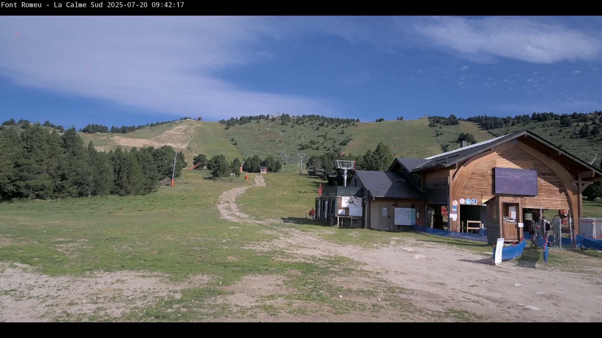 A grassy mountainside with a ski lift and a wooden building is visible under a mostly sunny sky with a few clouds.