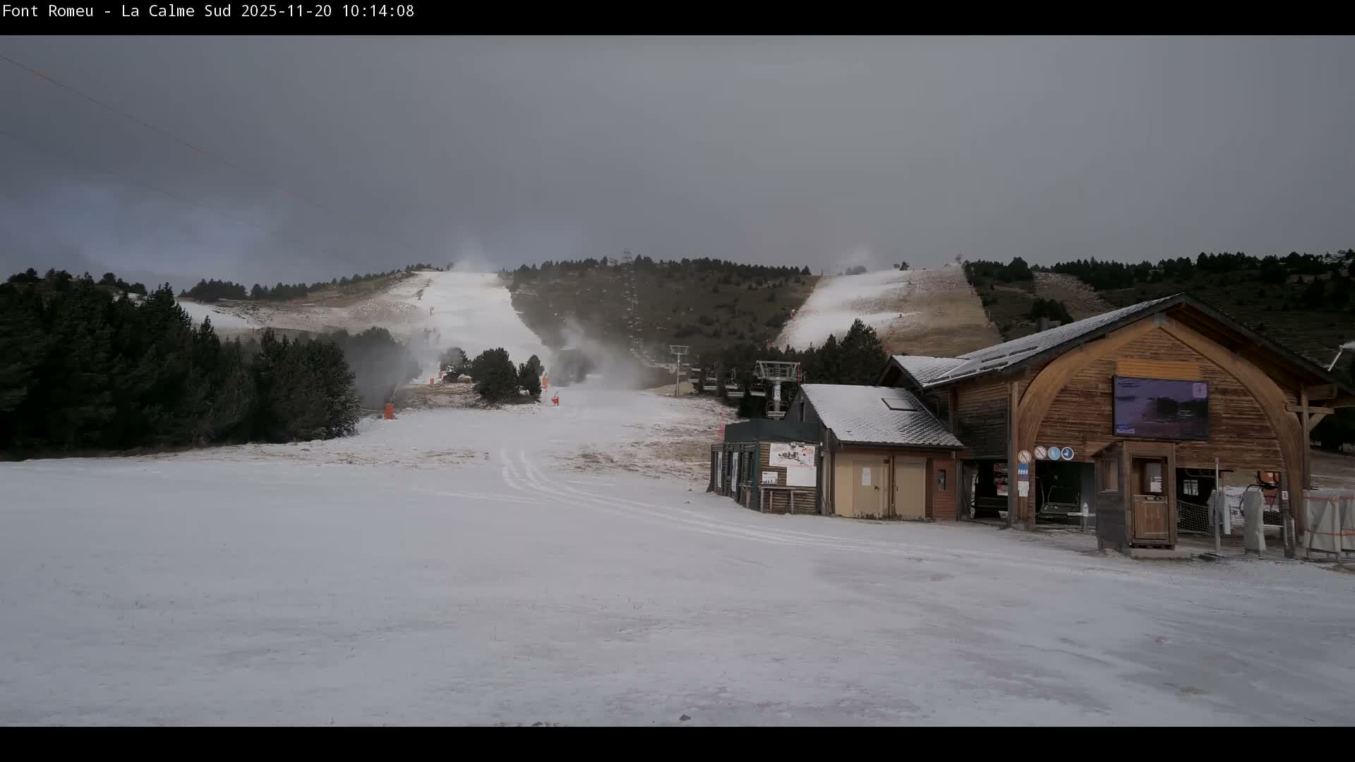 A winter ski resort scene displays partially snow-covered slopes with active snow cannons, ski lifts, evergreen trees, and wooden buildings, all beneath a grey, overcast sky.