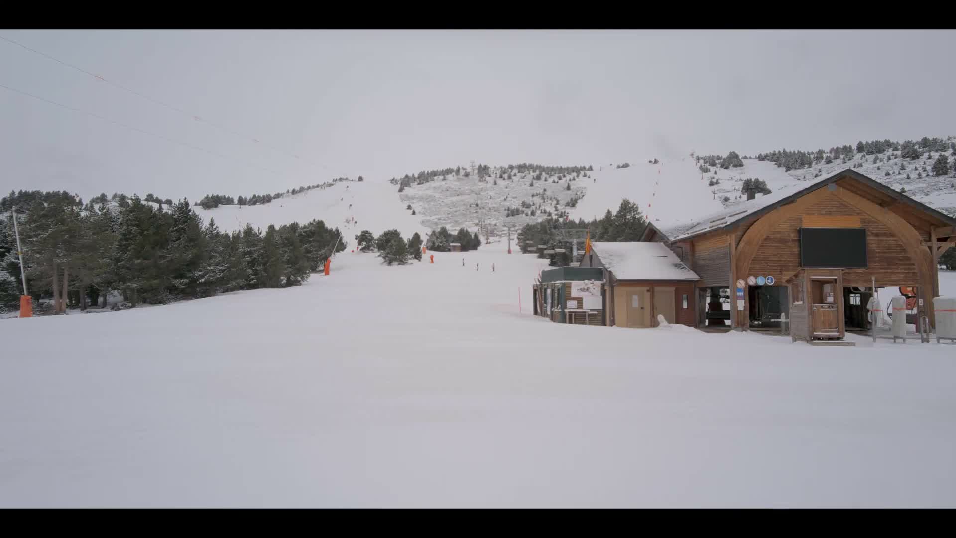 A winter ski resort scene displays partially snow-covered slopes with active snow cannons, ski lifts, evergreen trees, and wooden buildings, all beneath a grey, overcast sky.