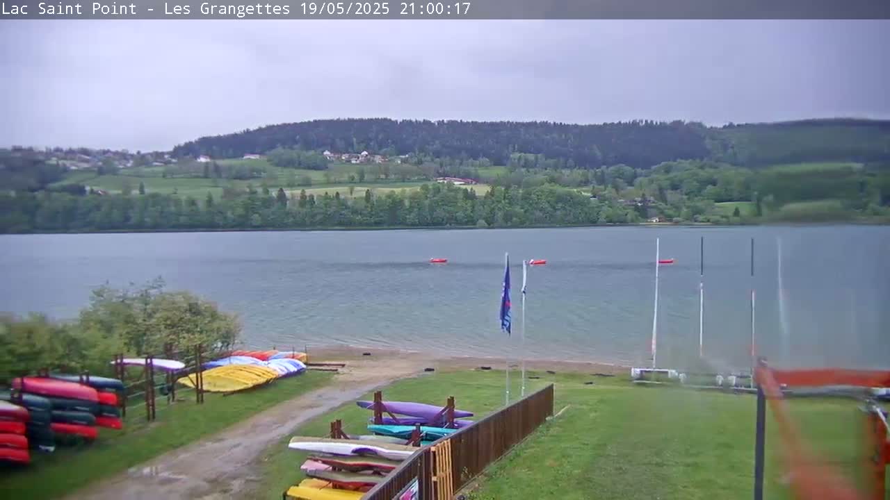 A calm lake under an overcast sky is surrounded by green hills and trees, with several boats and kayaks stored near the shoreline.