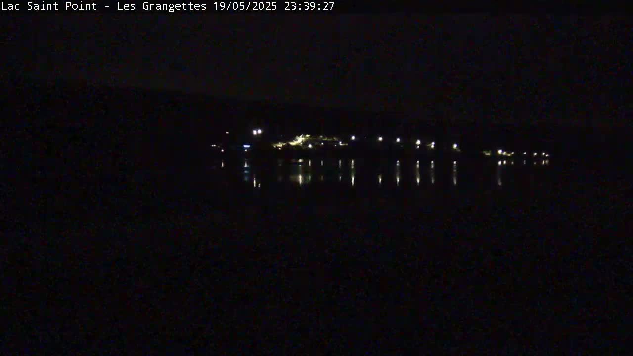Lights from a town across a dark body of water are reflected in the still water under a nighttime sky.