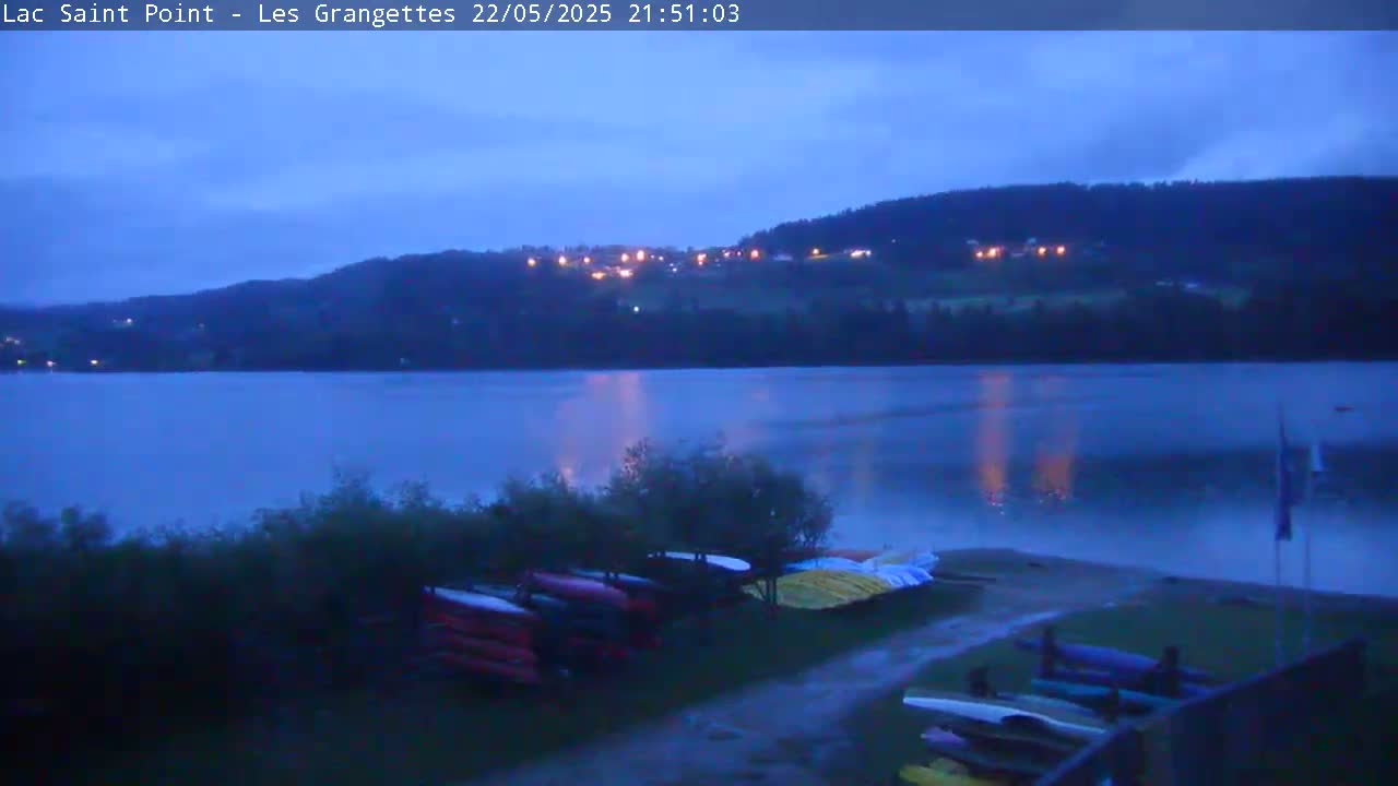 Under a twilight sky, a calm lake reflects the lights of a distant hillside village, with several canoes stored on the shore.