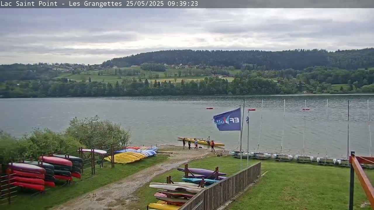 A calm lake is surrounded by green hills under an overcast sky, with several canoes and sailboats docked near a shoreline where several people are gathered.
