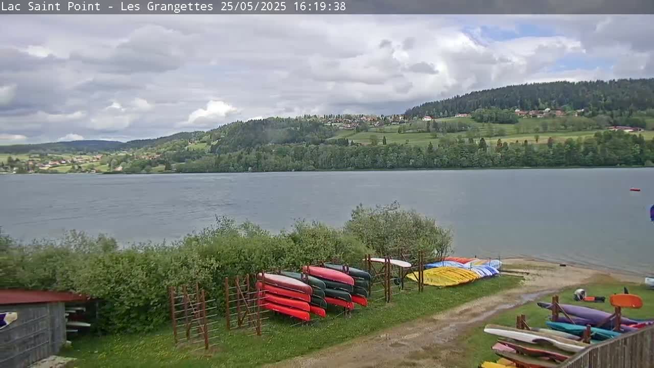 Canoes are stored near a calm lake under a partly cloudy sky, with a village and forest visible in the distance.