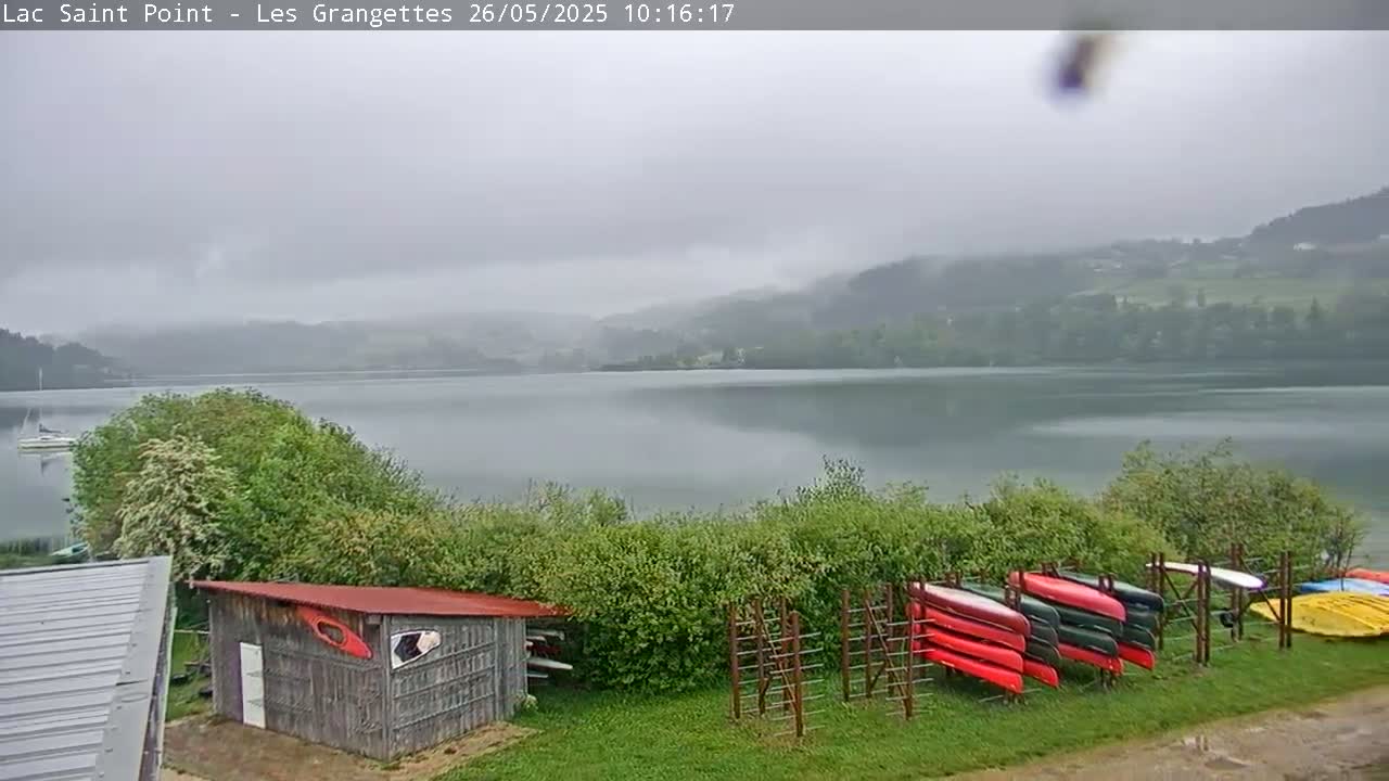 A calm lake under an overcast sky is viewed from a lakeside vantage point showing several canoes stored upright on racks near a small shed.