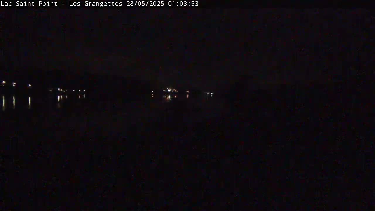 A dark nighttime scene shows a body of water reflecting faint lights from structures on the opposite shore.