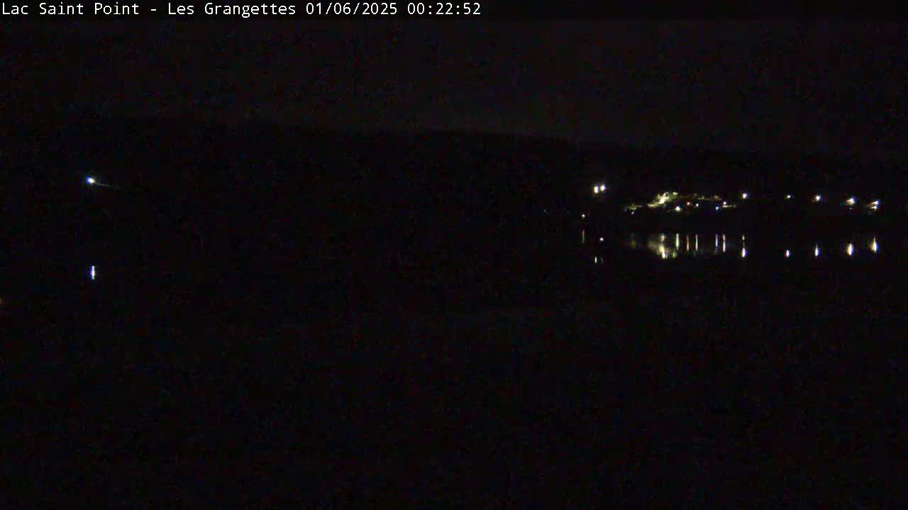 A nighttime view shows a dark body of water reflecting sparse lights from a distant shoreline under a clear, dark sky.