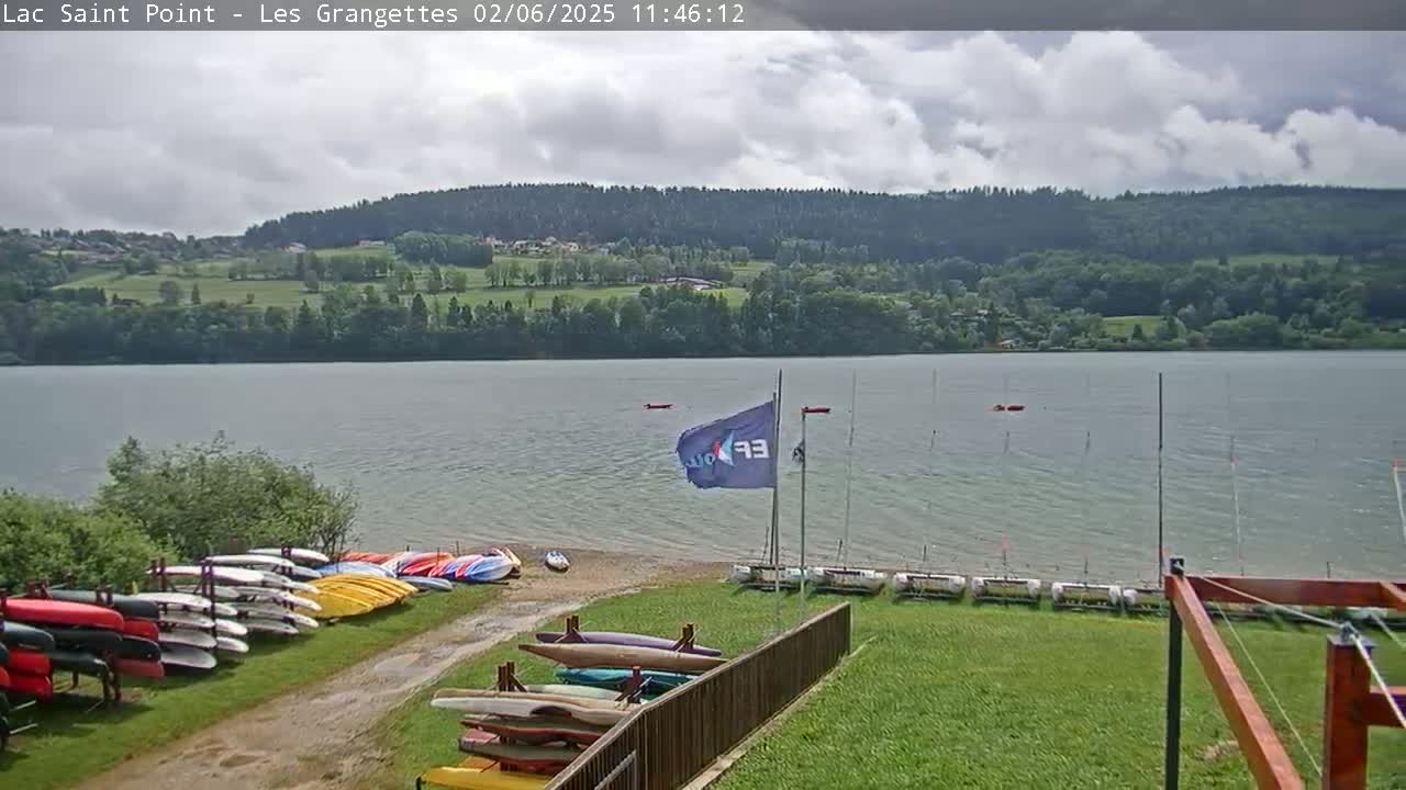 A calm lake with a partly cloudy sky, numerous kayaks and sailboats on the shore, and a forested hillside in the background.