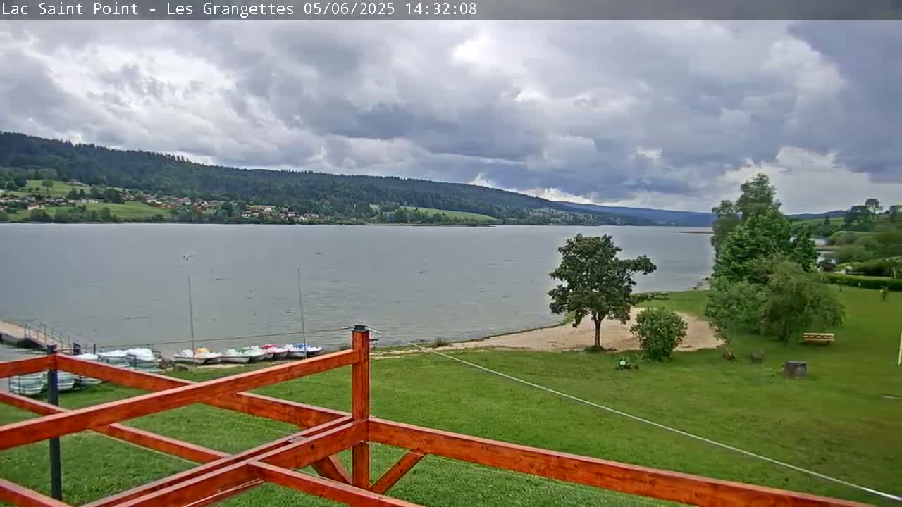 A partly cloudy day overlooks a calm lake with a grassy shoreline, several small boats docked, and a wooden structure in the foreground.