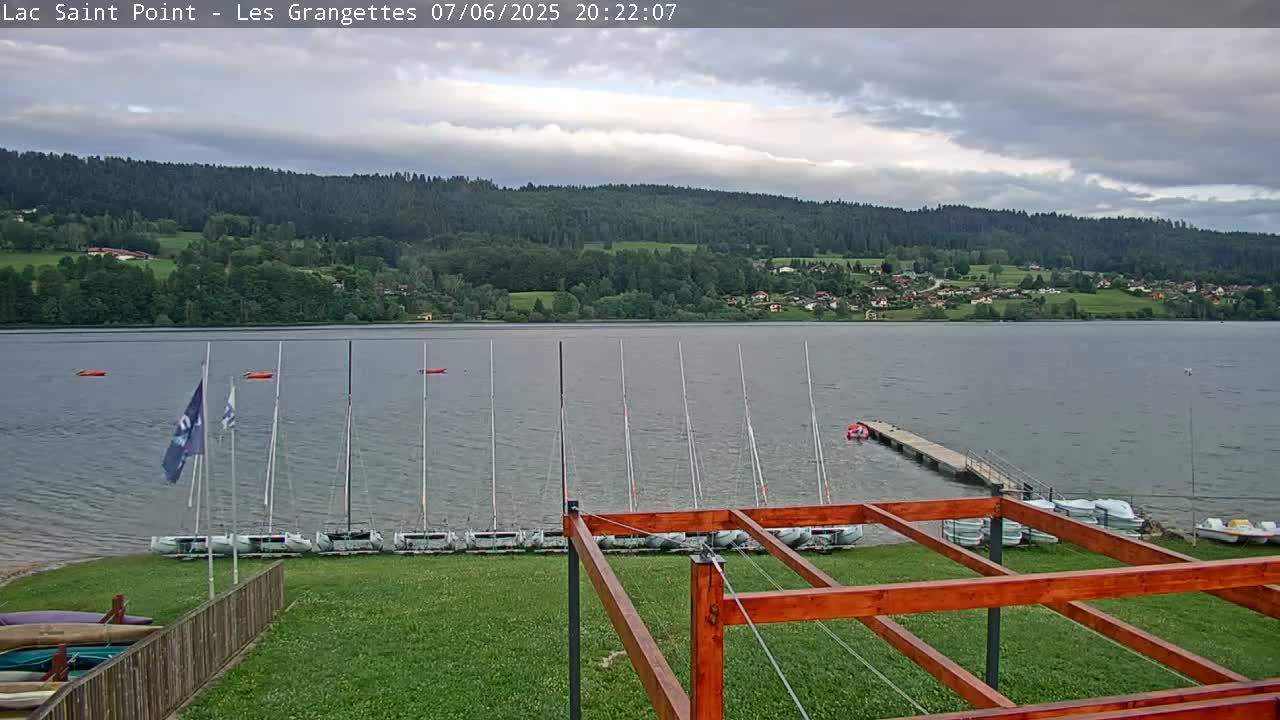 Several sailboats are docked at a lakeside area under a partly cloudy sky, with a forested hill in the background.