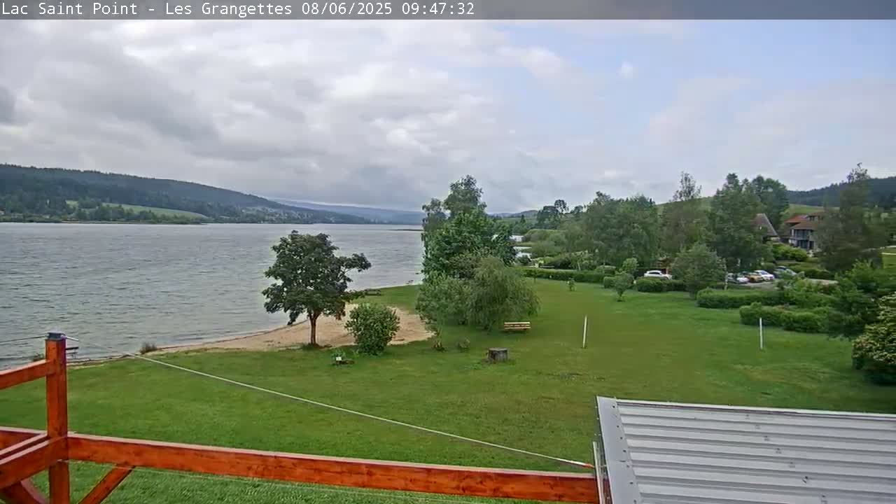 A calm lake stretches out under a partly cloudy sky, bordered by green grassy areas with trees and a small sandy beach, all viewed from a wooden deck.