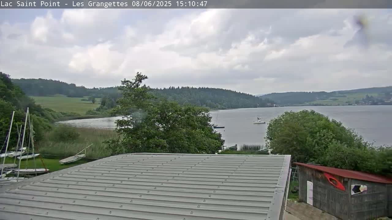 A mostly cloudy day overlooks a calm lake with several sailboats, surrounded by green trees and fields, from a rooftop with a corrugated metal surface.