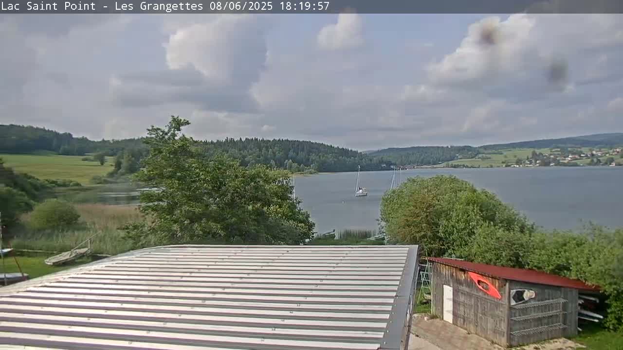 A partly cloudy day overlooks a calm lake with sailboats, bordered by green trees and fields, and a corrugated metal roof in the foreground.